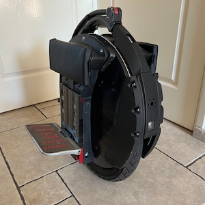 Op de afbeelding: Een zwarte elektrische eenwieler met een rode handgreep en een voetensteun. De eenwieler heeft een zwarte gepolsterde stoel en een zwart wiel met een zilveren velg.