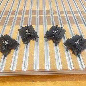 Può includere: Quattro morsetti in plastica nera con viti in metallo, montati su una superficie in legno con strisce di metallo argentate.