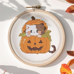 May include: A cross-stitch embroidery hoop featuring a cat in a pumpkin design. The cat has white and brown fur. The pumpkin is orange with a carved face. The hoop is surrounded by fall foliage with orange and brown leaves.