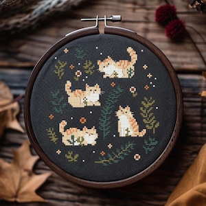 May include: A cross-stitch embroidery hoop featuring four orange and white tabby cats on a black background. The cats are surrounded by green foliage and small white dots. The wooden hoop is displayed on a wooden surface.
