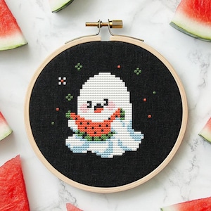 Könnte beinhalten: Ein weißer Geist mit freundlichem Gesicht hält eine Scheibe Wassermelone. Der Geist ist auf einem schwarzen Stoff gestickt, der über einen hölzernen Stickrahmen gespannt ist. Der Rahmen ist von Wassermelonenstücken umgeben.