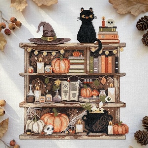 Peut inclure: Une broderie au point de croix sur tissu blanc représentant une étagère en bois à trois niveaux remplie d'objets d'Halloween. On y voit un chat noir, un chapeau de sorcière, des citrouilles, des livres, des bougies, un chaudron et des toiles.
