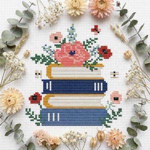 Pode incluir: Um design de ponto cruz com uma pilha de três livros com lombadas azuis e douradas, encimada por um buquê de flores rosa e vermelhas e folhagem verde. O design é cercado por flores secas e folhas de eucalipto.