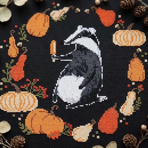 Peut inclure: Une broderie au point de croix sur tissu noir représentant un blaireau central tenant une glace à l'eau orange, entouré d'un arrangement circulaire de diverses citrouilles et courges orange. Le motif utilise un style pixelisé aux tons vifs.