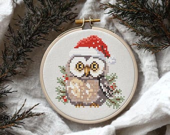 May include: A cross-stitch pattern of an owl wearing a red Santa hat with green holly and red berries. The owl is stitched on a white fabric and framed in a wooden hoop.