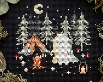 May include: A cross-stitch embroidery on black fabric featuring a white ghost camping in a forest. The design includes a tent, a campfire with a roasting stick, a lantern, small red mushrooms, a crescent moon, and stars in shades of white and gray.