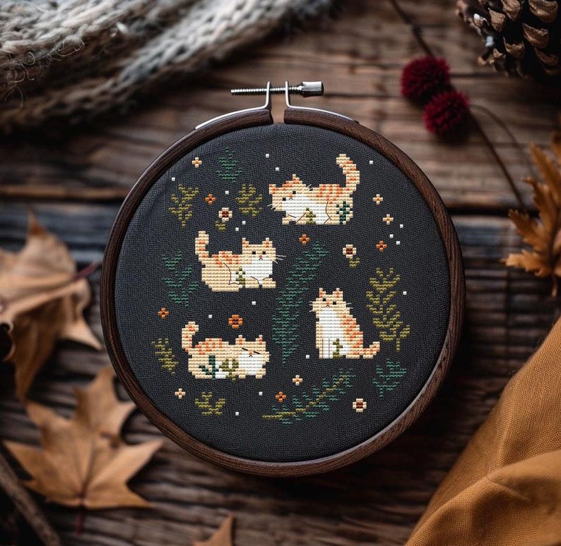 May include: A cross-stitch embroidery hoop featuring four orange and white tabby cats on a black background. The cats are surrounded by green foliage and small white dots. The wooden hoop is displayed on a wooden surface.