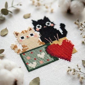 May include: Cross-stitch design featuring two cats and a red heart. One cat is black, the other is tan and orange. They are positioned above a green box with white and pink heart accents. The design is on a white fabric background.