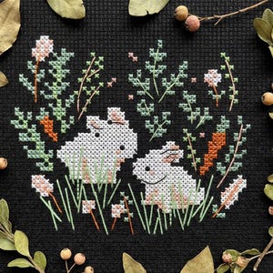 Op de afbeelding: Kruissteekborduurwerk op zwarte stof met twee witte konijnen omringd door groene bladeren, oranje wortels en roze bloemen. Het ontwerp is omlijst door gedroogde bladeren en bessen.