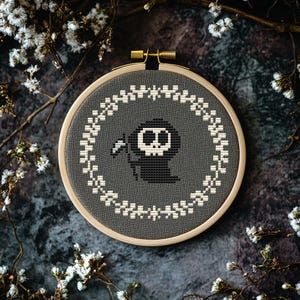 May include: A gray embroidery hoop with a black and white cross-stitch design of a cartoon Grim Reaper holding a scythe. The design is surrounded by a white wreath.