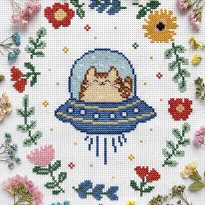 Könnte beinhalten: Kreuzstichmuster mit einer lächelnden Katze in einer blauen UFO, umgeben von einem floralen Kranz aus roten, rosa, gelben und grünen Blumen und Blättern. Die Katze trägt einen blauen Weltraumhelm. Der Hintergrund ist weiß.