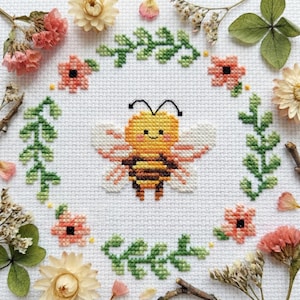 May include: A cross-stitch design of a smiling bee with yellow and brown stripes, encircled by a green leaf border and small pink flowers. The design is set against a white background and accented with dried flowers.