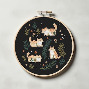 May include: A cross-stitch embroidery hoop featuring four orange and white cats in various poses, surrounded by green foliage and small orange flowers, set against a black background. The hoop is made of wood.