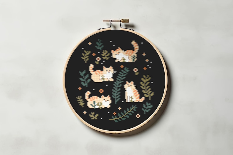 May include: A cross-stitch embroidery hoop featuring four orange and white cats in various poses, surrounded by green foliage and small orange flowers, set against a black background. The hoop is made of wood.