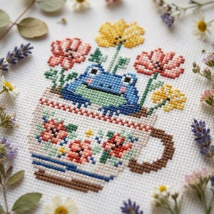 Può includere: Ricamo a punto croce con una rana blu in una tazza floreale. La tazza è decorata con fiori nei toni del rosso, rosa e giallo. La rana è circondata da fiori, creando una scena stravagante e affascinante.
