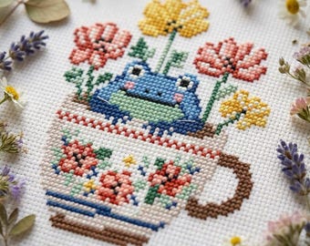 May include: Cross-stitch art featuring a blue frog in a floral teacup. The teacup is decorated with flowers in shades of red, pink, and yellow. The frog is surrounded by flowers, creating a whimsical and charming scene.