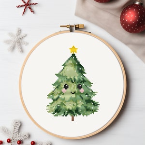 May include: A cross-stitch embroidery hoop with a green Christmas tree with a yellow star on top. The tree has a smiling face with pink cheeks.
