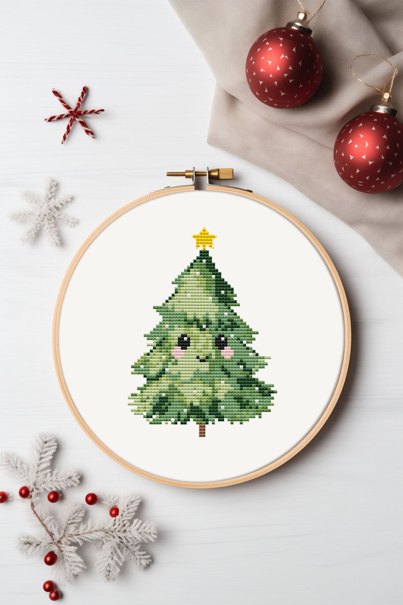 May include: A cross-stitch embroidery hoop with a green Christmas tree with a yellow star on top. The tree has a smiling face with pink cheeks.