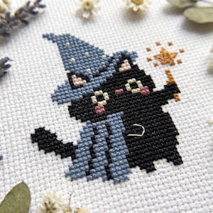 Peut inclure: Une broderie au point de croix sur tissu blanc représentant un chat noir portant un chapeau et une cape de magicien bleus. Le chat tient une petite baguette étoilée dorée. Le motif inclut des joues roses et des étoiles jaunes sur le chapeau.