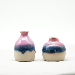Könnte beinhalten: Zwei Keramikvasen mit Farbverlauf. Die obere Hälfte jeder Vase ist eine Mischung aus Rosa und Blau, mit einem dunkelblauen Tropfeffekt. Die untere Hälfte ist naturfarben beige. Eine Vase ist hoch und schlank, die andere ist rund.