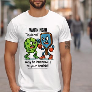 May include: White t-shirt with a cartoon graphic of a pickleball and paddle in a boxing stance. The text "WARNING!!! Pickleball may be hazardous to your health!!!" is printed below the graphic.