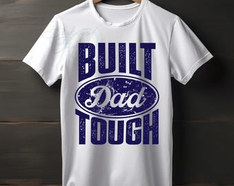 Puede incluir: Camiseta blanca con el texto "BUILT Dad TOUGH" en letras azul marino desgastadas. La palabra "Dad" está dentro de un óvalo. La camiseta está colgada en una percha de madera sobre un fondo de madera oscura.