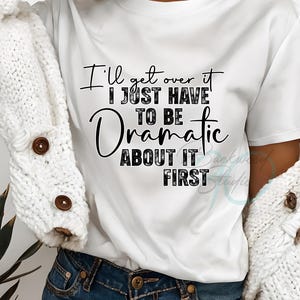 Puede incluir: Camiseta blanca con el texto "I'll get over it, I just have to be dramatic about it first" en escritura negra. La camiseta se combina con pantalones cortos de mezclilla azules. Un cárdigan blanco está sobre los hombros.