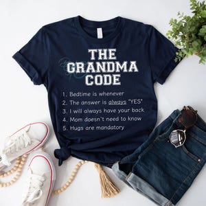 May include: A navy blue t-shirt with the words "THE GRANDMA CODE" in white. The shirt lists grandma rules. White trainers, denim shorts, and sunglasses are also visible in the image.