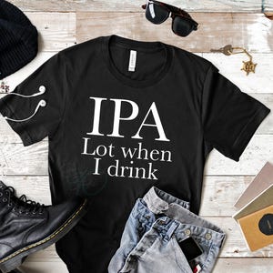 Può includere: T-shirt nero con la scritta bianca "IPA Lot when I drink". La maglietta è esposta con jeans blu, stivali neri, un berretto nero, occhiali da sole e altri accessori su una superficie di legno.