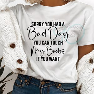 May include: White t-shirt with the text "SORRY YOU HAD A Bad Day YOU CAN TOUCH My Boobs IF YOU WANT" in black script. The shirt is worn with blue denim shorts and a white cardigan.