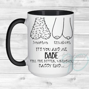 May include: White ceramic mug with a black handle and interior. The mug features a humorous illustration of two pairs of testicles and the text "It's You and Me Babe Till the Bitter, Wrinkly, Saggy End..." with the names "Jonathon" and "Elizabeth".