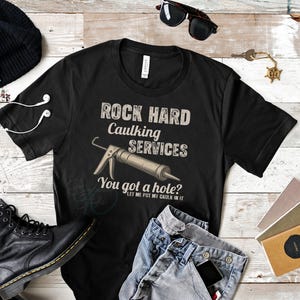 Peut inclure: T-shirt noir avec le texte "ROCK HARD Caulking SERVICES. You got a hole? LET ME PUT MY CAULK IN IT" et un dessin d'un pistolet à calfeutrer. Le t-shirt est présenté avec un jean, des bottes et des accessoires.