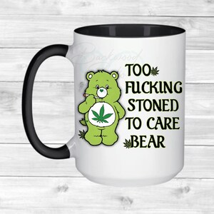 May include: White ceramic mug with a black handle and interior. The mug features a green bear smoking a cigarette with a marijuana leaf on its belly. Text on the mug reads "TOO FUCKING STONED TO CARE BEAR."