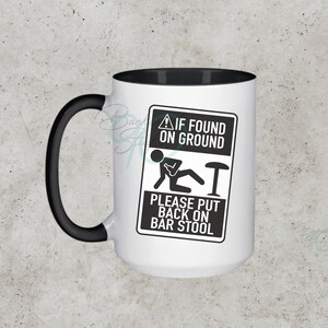 Può includere: Tazza in ceramica bianca con interno e manico neri, con un cartello umoristico: "If Found On Ground, Please Put Back On Bar Stool". La tazza è di dimensioni e forma standard, perfetta per caffè o tè.