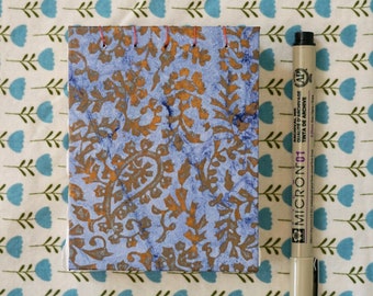 May include: A small blue and gold patterned notebook with a black and brown Micron 01 archival ink pen. The notebook is bound with a red thread and has a simple design.