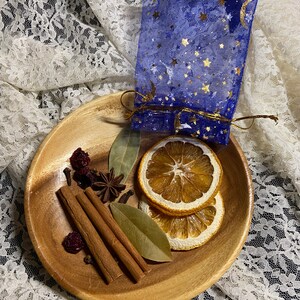 May include: A wooden bowl filled with dried orange slices, cinnamon sticks, bay leaves, star anise, and berries. A blue mesh bag with gold stars and moons is tied with a gold string. The items are arranged on a white lace surface.