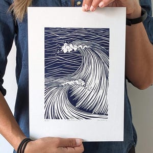 May include: A framed print with a navy blue and white wave design. The artwork shows a cresting wave with detailed linework, producing a dynamic and textured appearance. The print is held by a person.