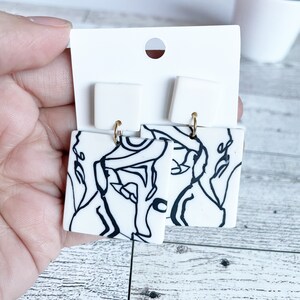 May include: A pair of white square earrings with black abstract line designs. The earrings have gold hooks.