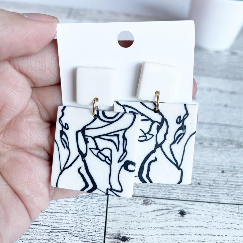 May include: A pair of white square earrings with black abstract line designs. The earrings have gold hooks.