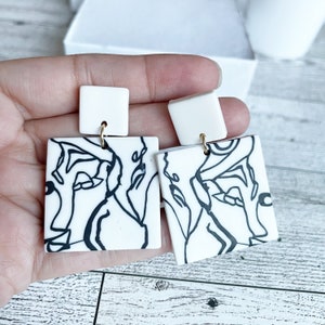 May include: A pair of white square earrings with black line art designs. The earrings have gold hooks.