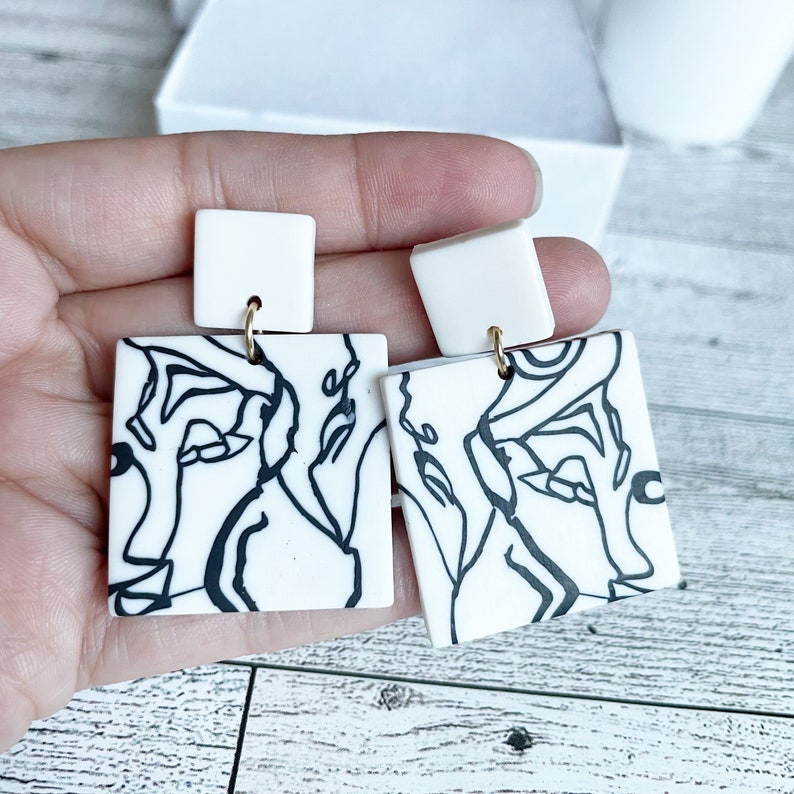 May include: A pair of white square earrings with black line art designs. The earrings have gold hooks.
