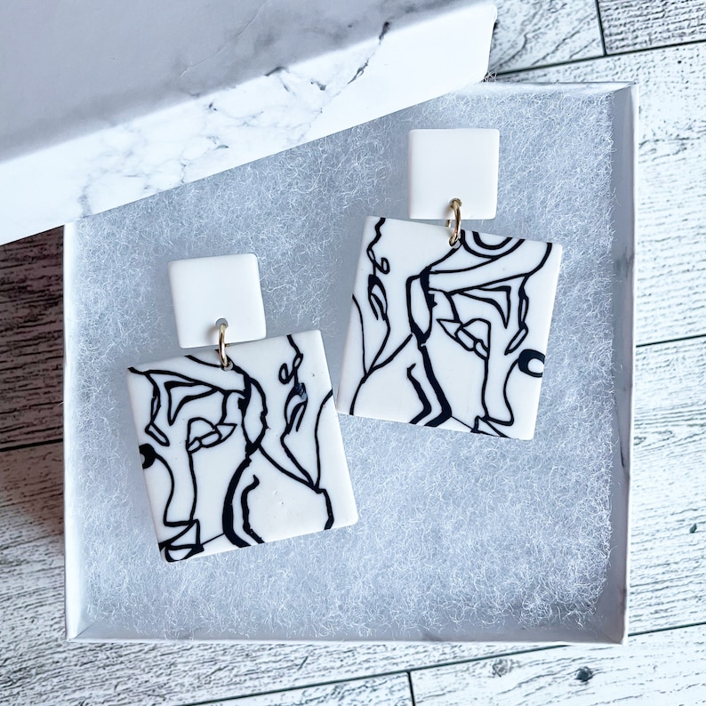 May include: A pair of white square earrings with black line drawings of abstract faces. The earrings have gold hooks.