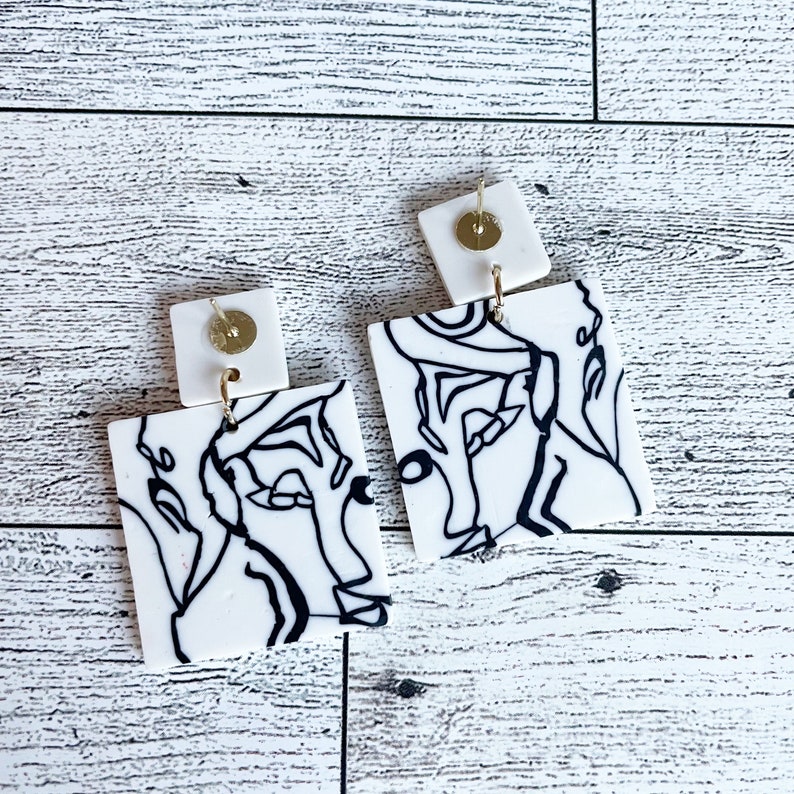 May include: A pair of white square earrings with black line drawings of abstract faces. The earrings have gold posts and backs.