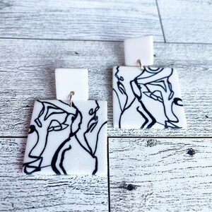 May include: A pair of white square earrings with black line art designs. The earrings have gold hooks.