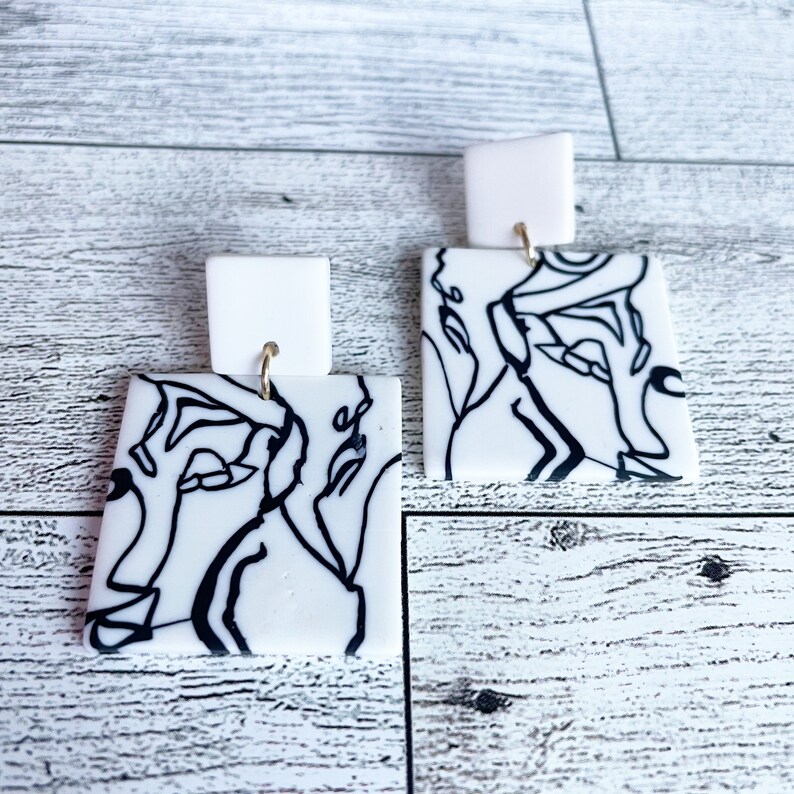 May include: A pair of white square earrings with black line art designs. The earrings have gold hooks.