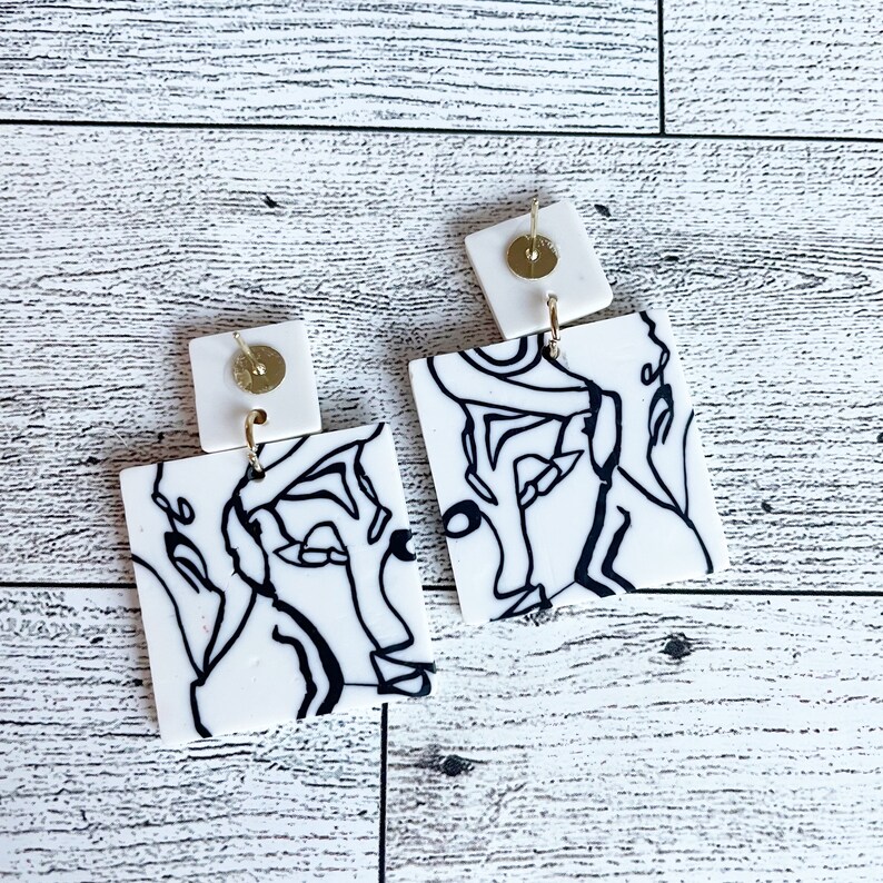 May include: A pair of white square earrings with black line drawings of abstract faces. The earrings have gold posts and backs.