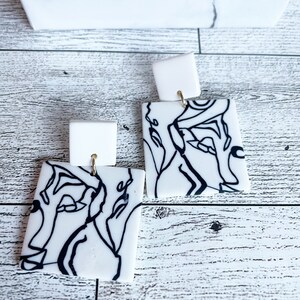 May include: A pair of white square earrings with black line drawings of abstract faces. The earrings have gold hooks.