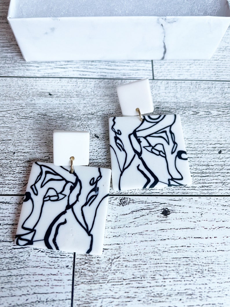 May include: A pair of white square earrings with black line drawings of abstract faces. The earrings have gold hooks.