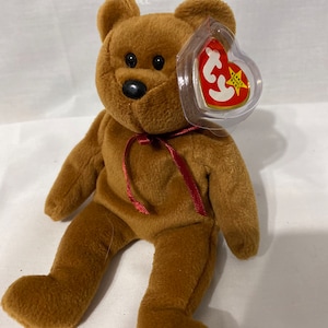 May include: A brown plush teddy bear with a red ribbon around its neck. The bear has a black nose and black eyes. The bear is wearing a Ty Beanie Baby tag with a white star and the words "Beanie Baby" on it.