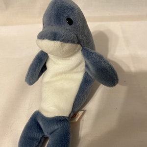 May include: A blue and white plush dolphin toy. The dolphin has a white belly and a blue back. The toy has a Ty tag attached to its right leg.